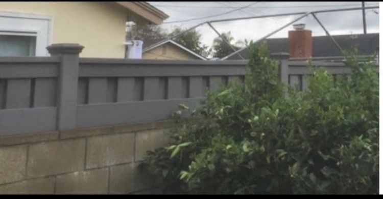 Gray wooden fence atop a concrete block wall, with green bush in the foreground, and a house in the background.