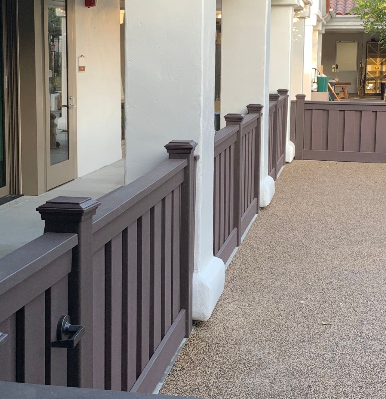 Brown wooden fence alongside a building with white pillars and a gravel surface.