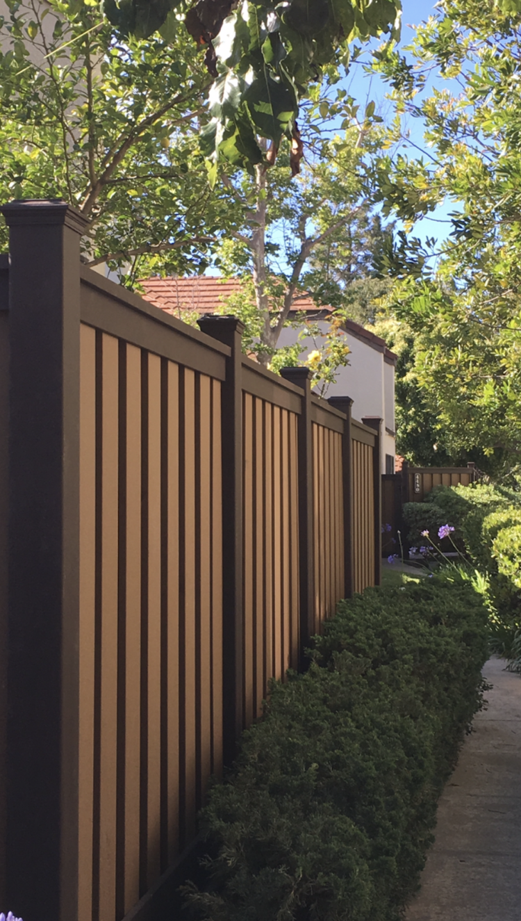 Brown and tan vertical fence with green bushes and trees in the background.
