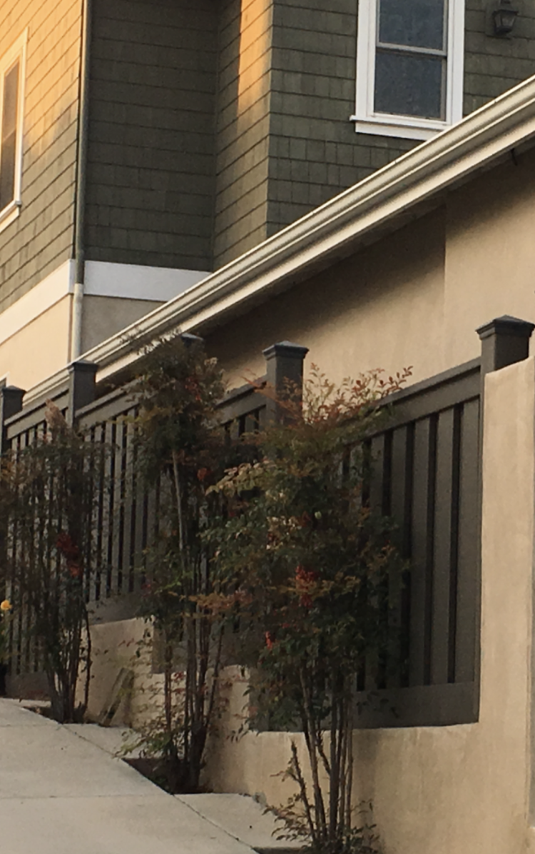 Dark fence with posts and shrubs in front of a building with green siding and a sidewalk.