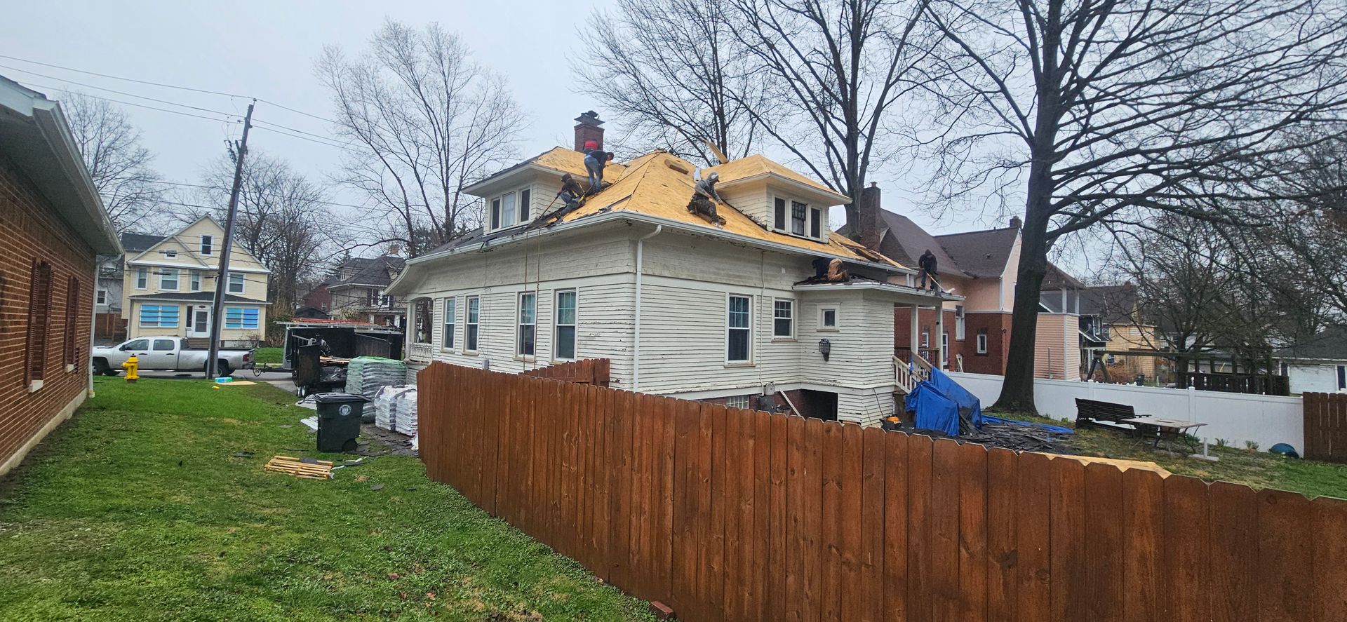 Rainy backyard with a white house, brown fence, bare trees, and a parked car in the distance.