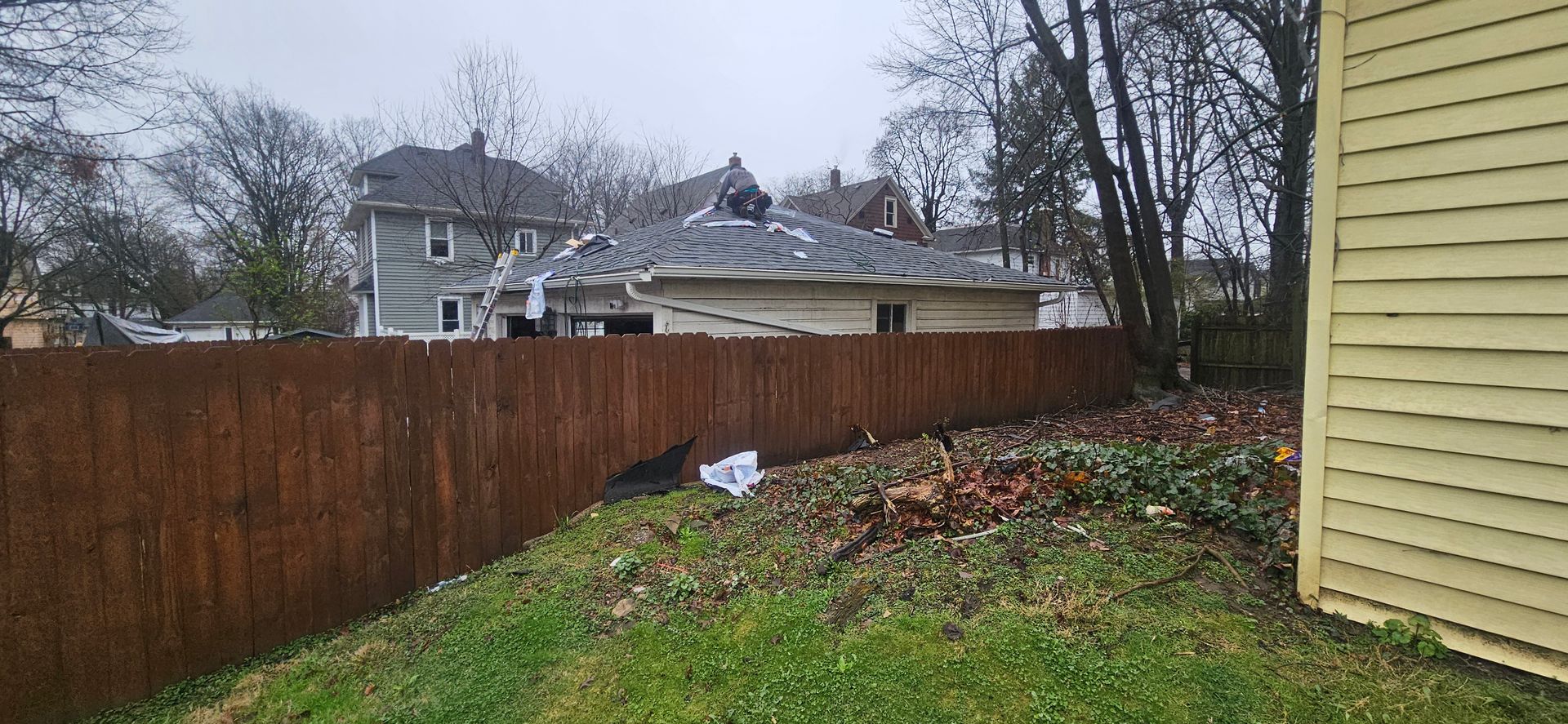 Rainy backyard with wooden fence, houses in the distance, and flower beds beside a yellow house