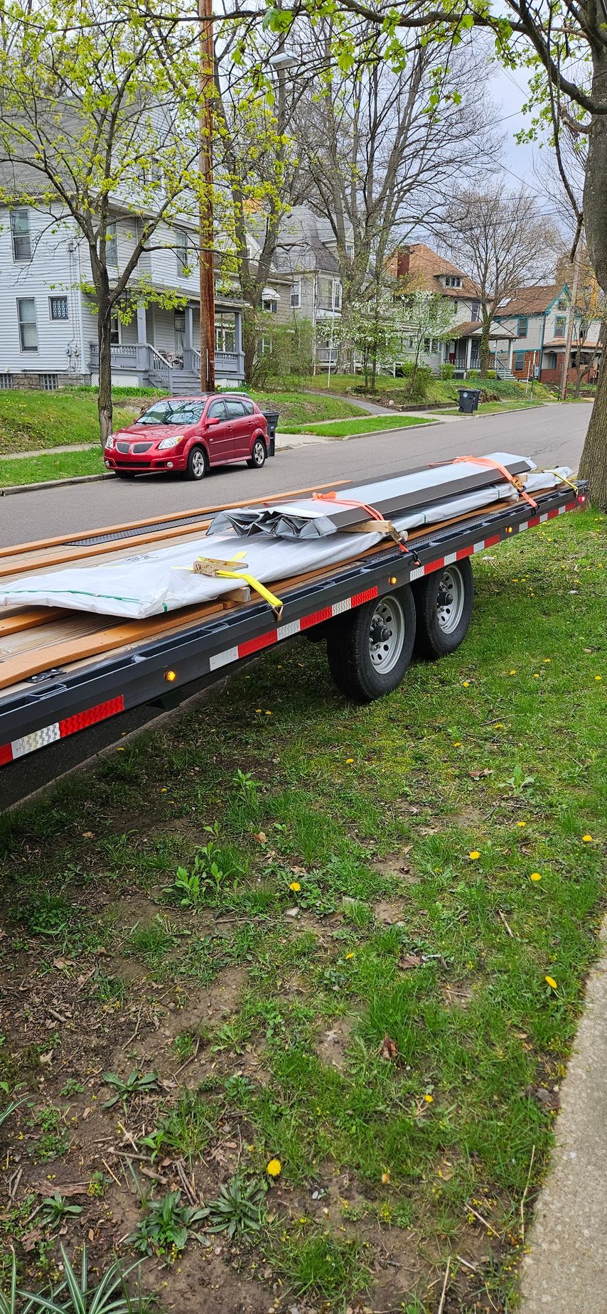 Flatbed trailer loaded with boards and metal panels parked on a grassy residential street.