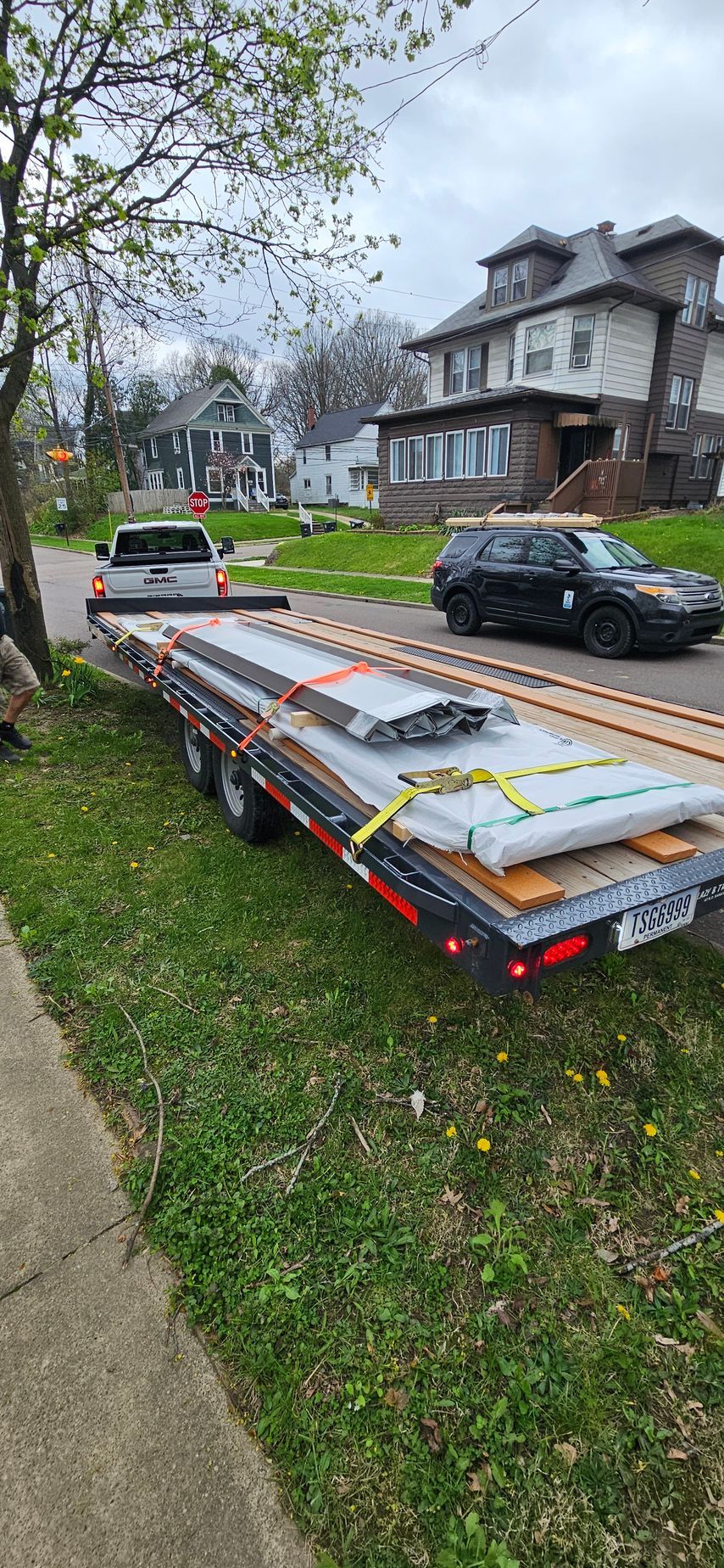 Flatbed trailer carrying construction materials parked on a grassy street beside houses and parked cars.