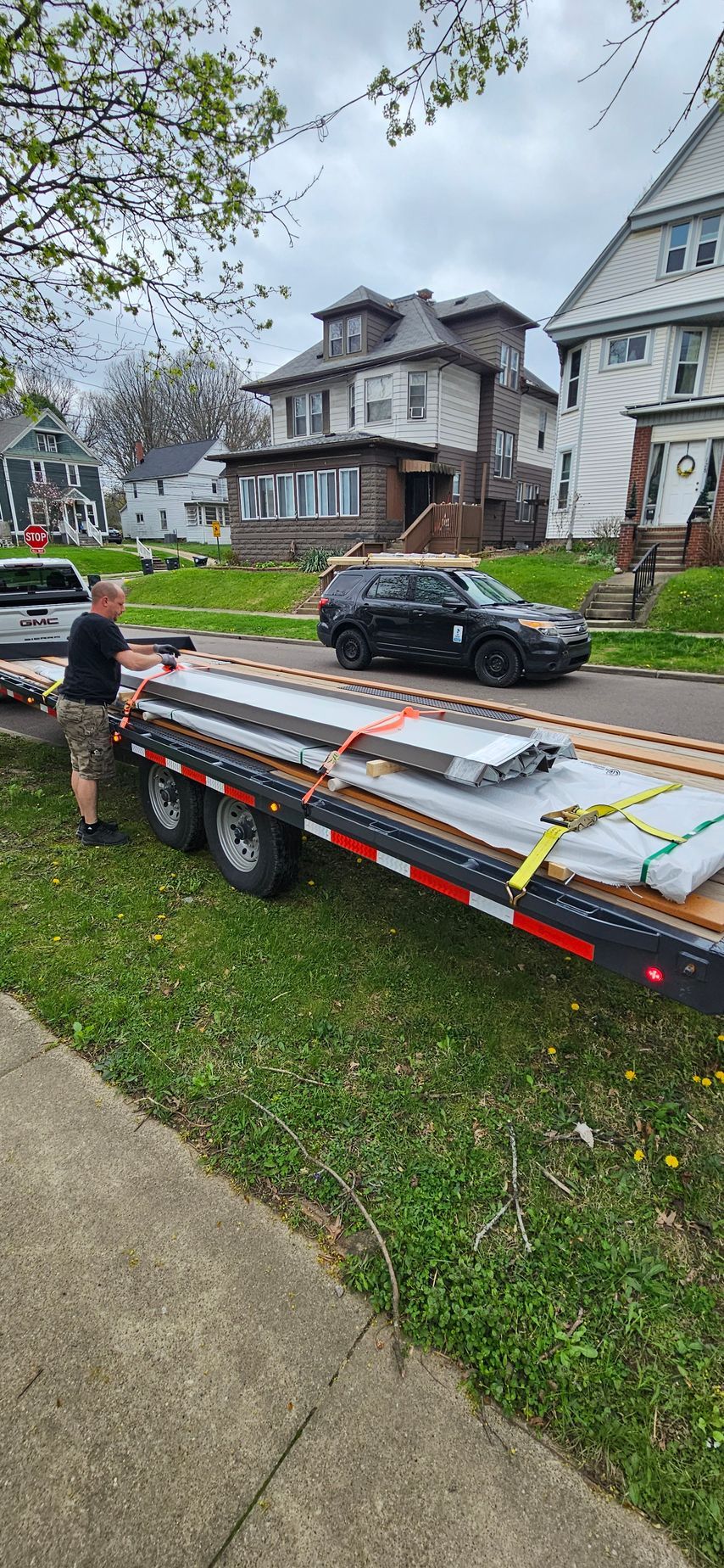 Flatbed trailer carrying long lumber on a residential street with houses and parked cars