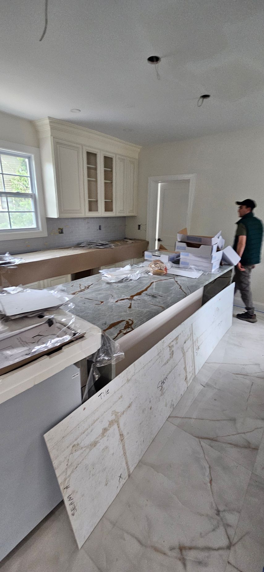Kitchen under renovation with white cabinets, marble countertops, and a worker installing the island
