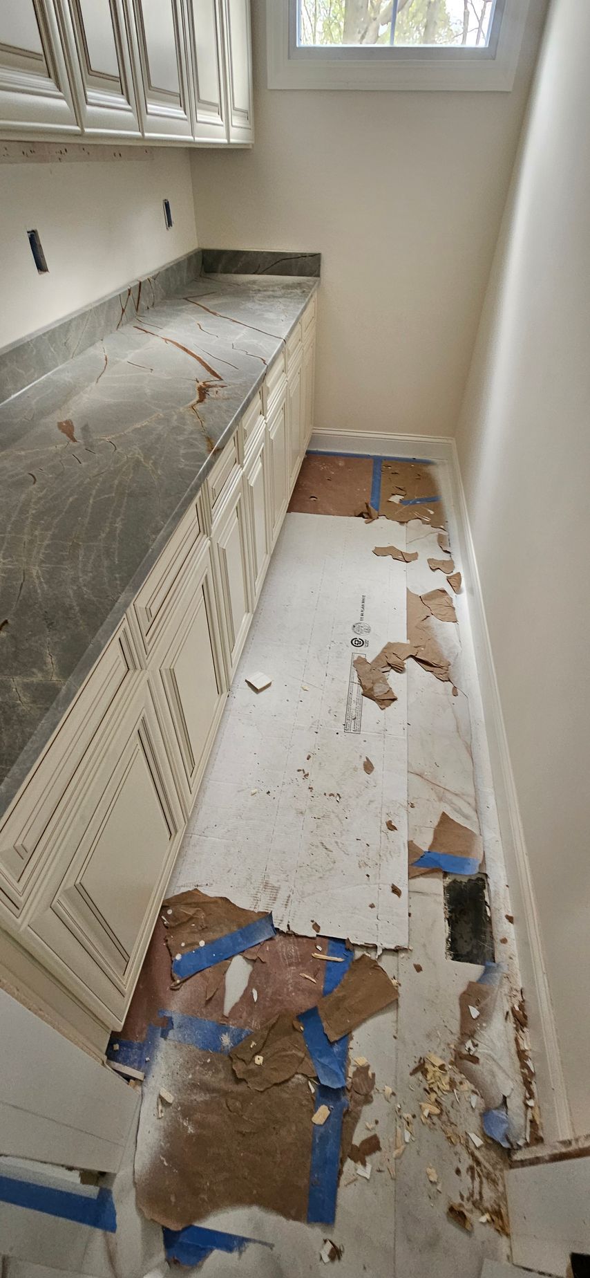 Damaged kitchen floor with torn linoleum, debris, and cabinets along one side near a window.