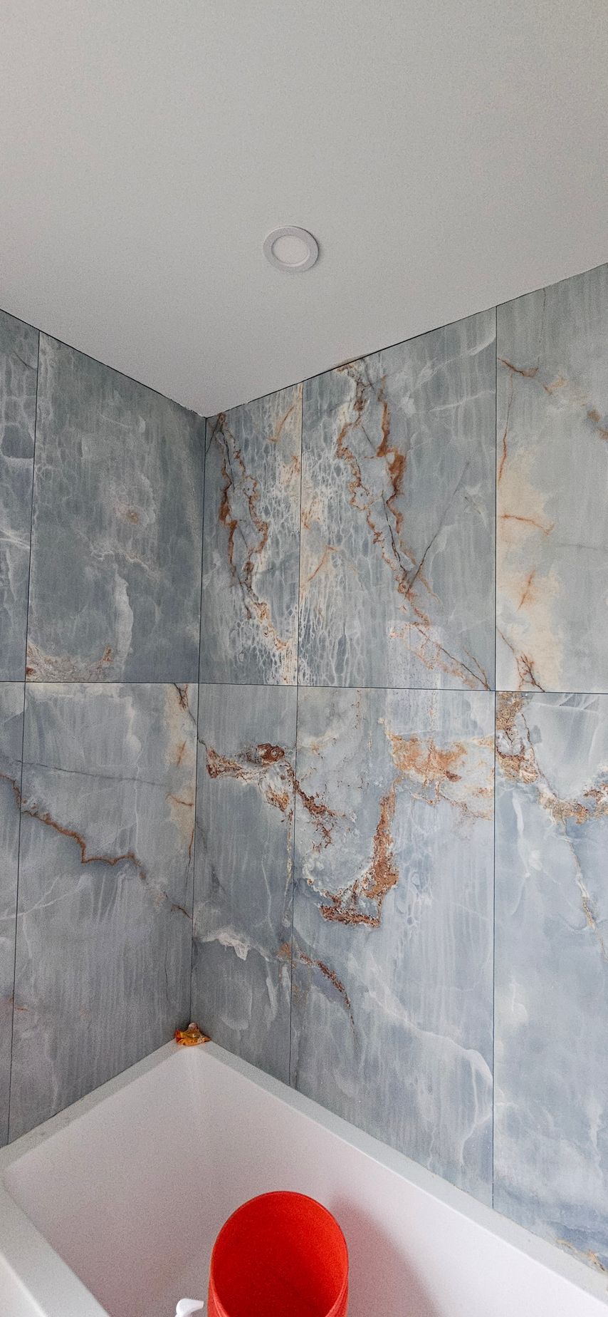 Modern shower corner with gray-blue marble tile and a red bucket in a white tub