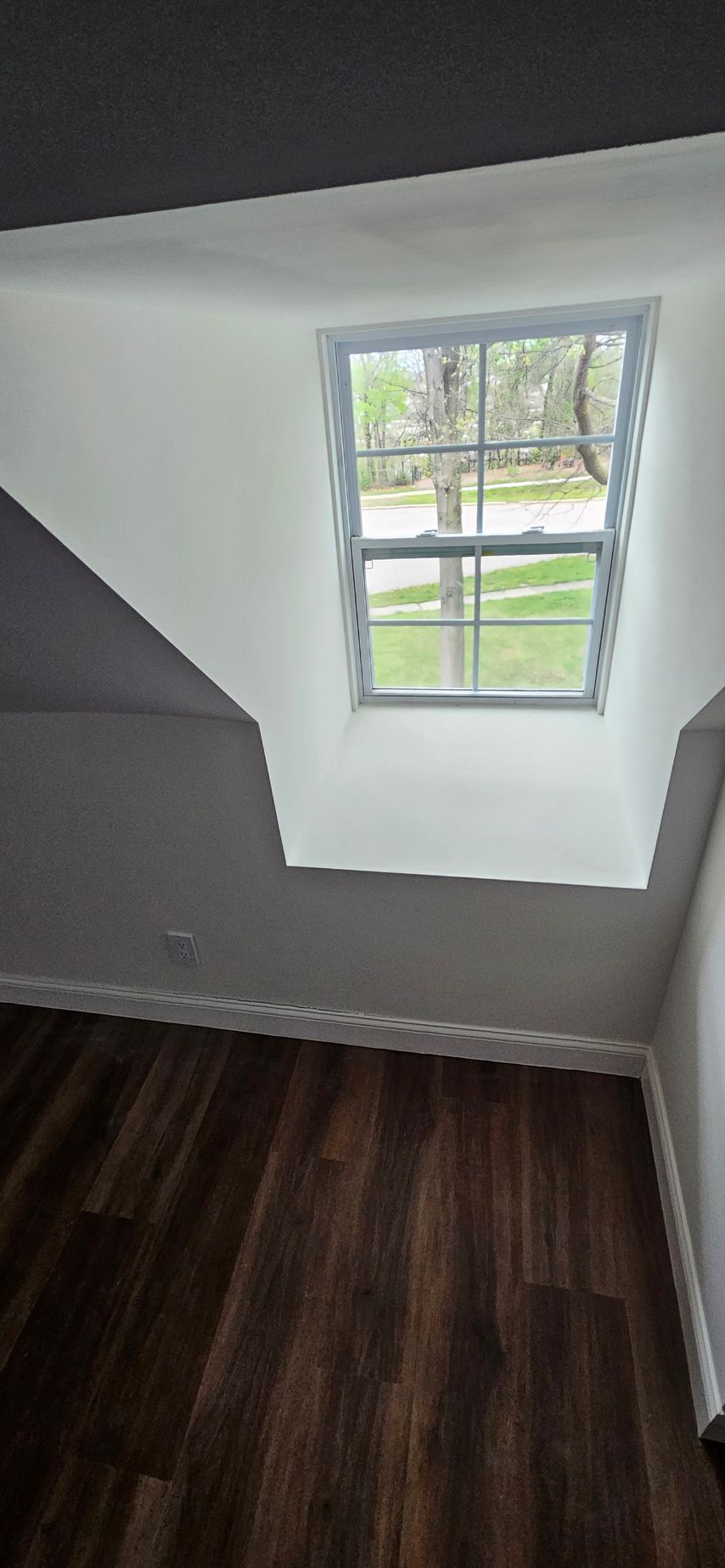 Small sloped room with a white-framed window, white walls, and dark wood flooring.