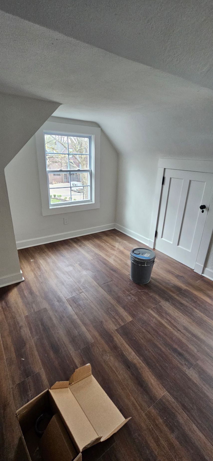 Empty attic room with sloped white ceiling, wood floors, window, closet, and moving boxes