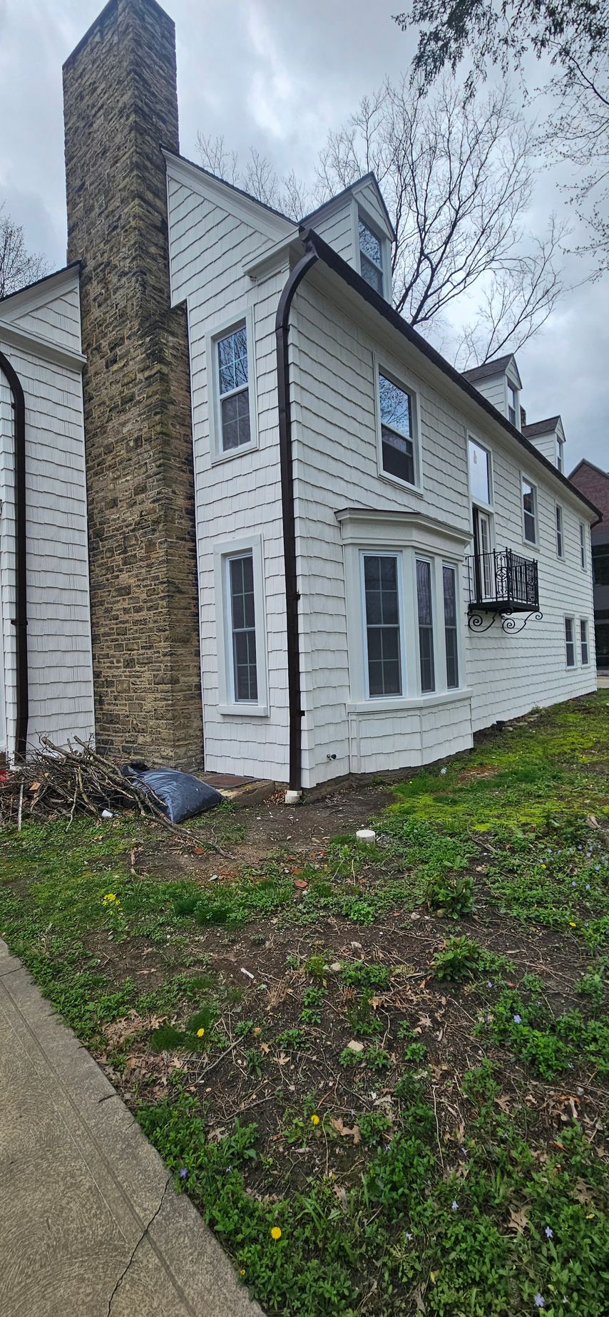 Two-story white house with stone chimney, black shutters, and a small leaf-covered yard beside a sidewalk.