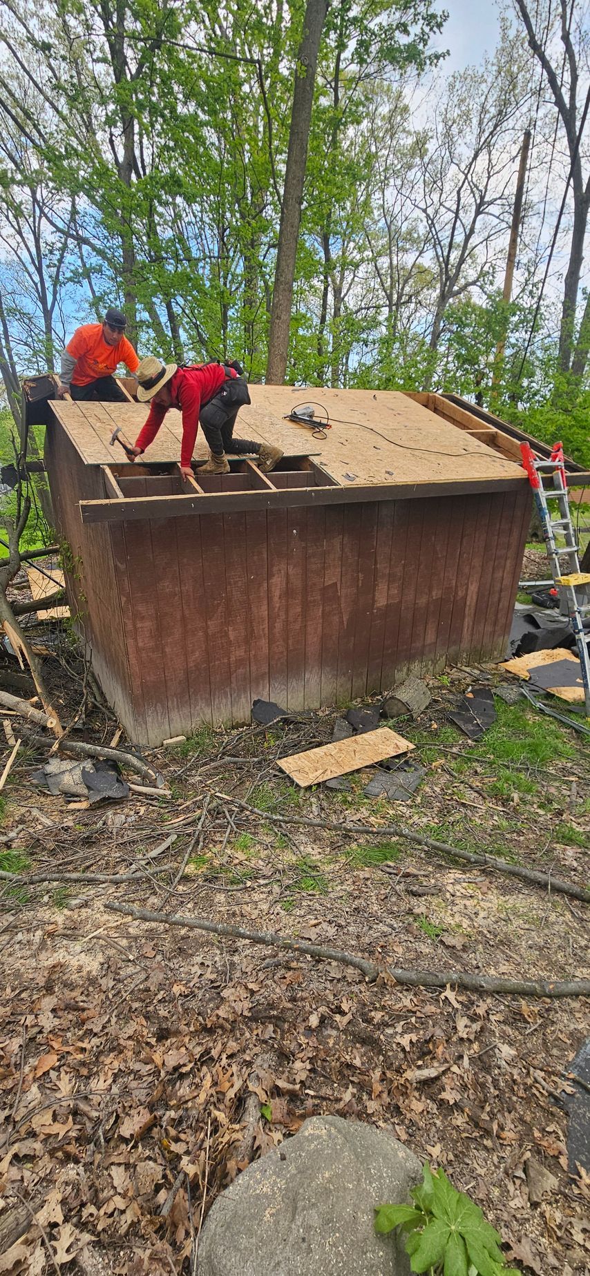 Two people in red shirts work on the roof of a rusted shed in a wooded area.