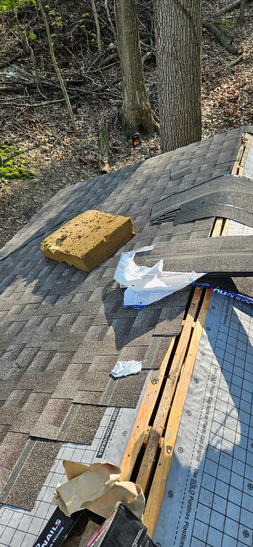 Roofing materials on a sloped roof beside trees, with yellow insulation, shingles, and tools visible.