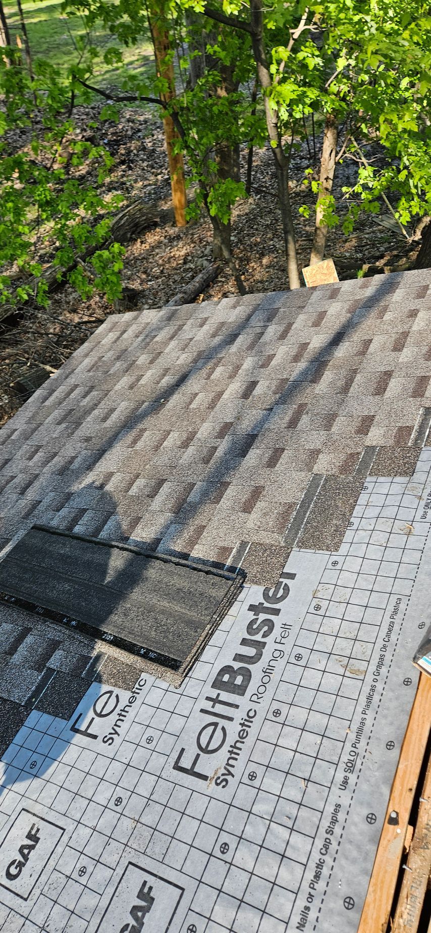 Roofer installing gray shingles over black underlayment beside leafy trees, with synthetic roof deck visible