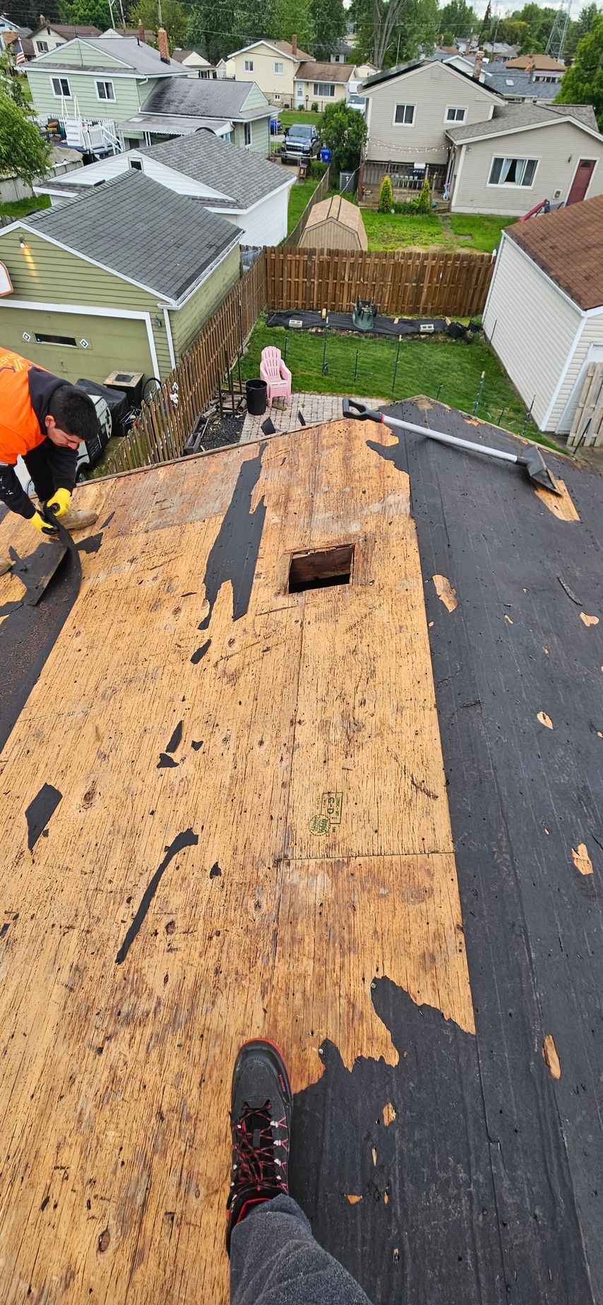 Roofer removing damaged shingles on a house roof, with plywood exposed and a residential neighborhood below.