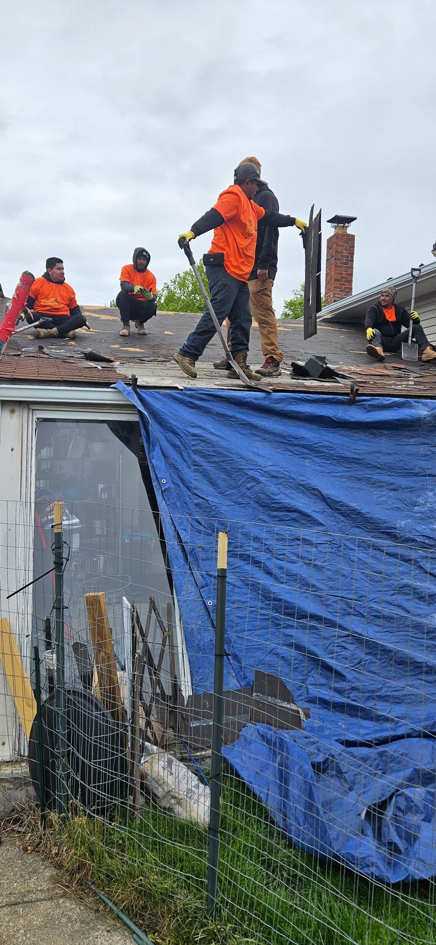 Workers in orange vests on a roof, standing over a blue tarp covering the building facade.