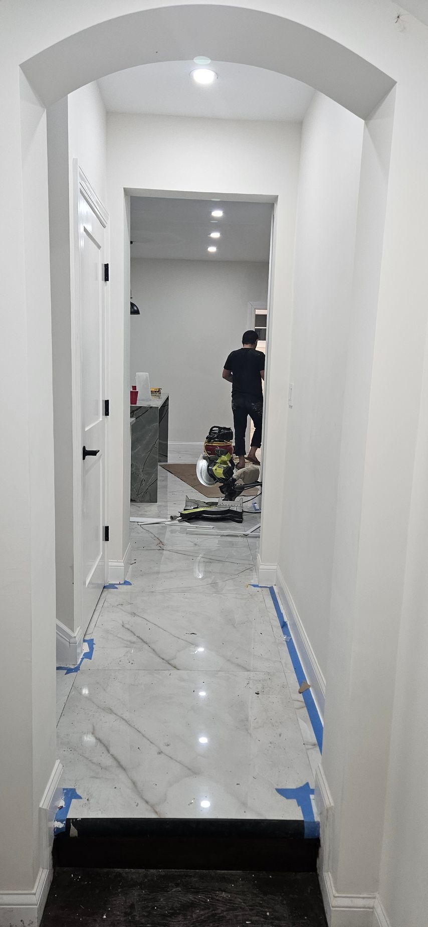 Narrow white hallway with marble floor, leading to a room where a person stands near cleaning supplies.