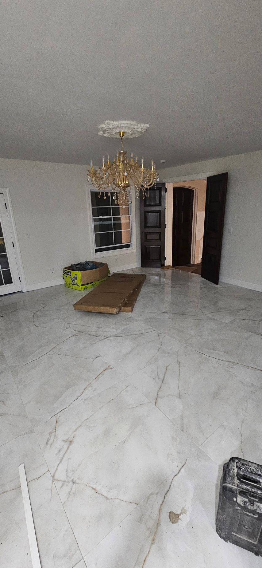 Empty living room with white tile floor, chandelier, and open doors leading to a hallway.
