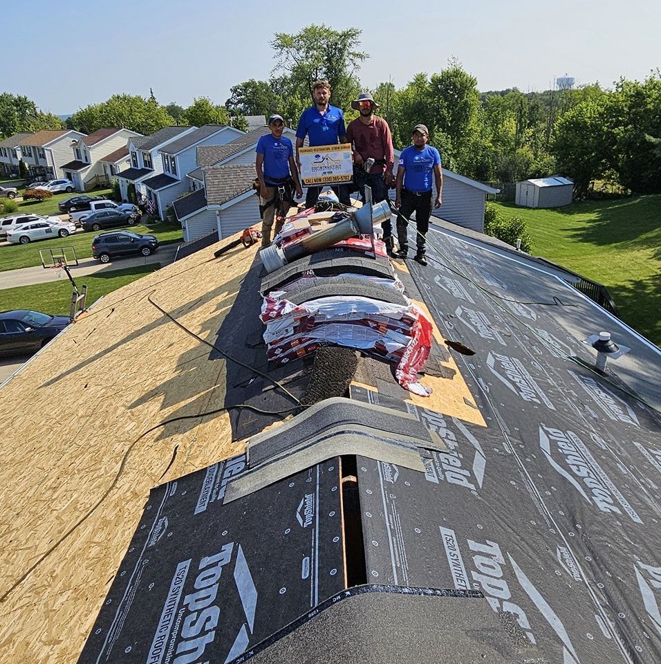 Four construction workers stand on a residential roof under repair, holding a promotional sign.