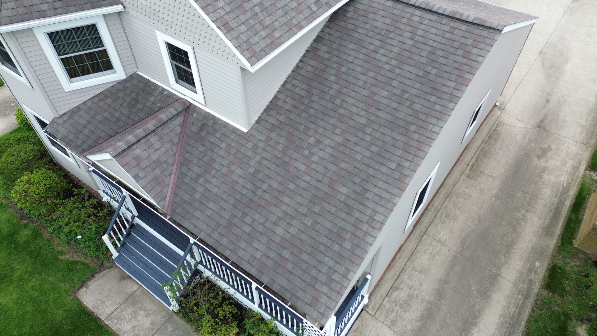 Aerial view of a gray house roof with white siding, dormer windows, and a small front porch by a driveway