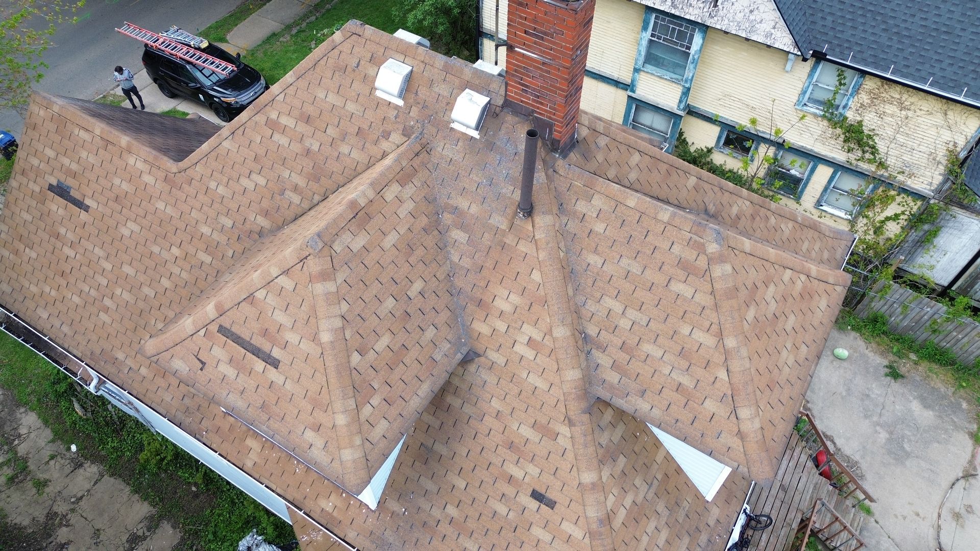 Aerial view of a brown shingled house roof with a brick chimney and surrounding yard.