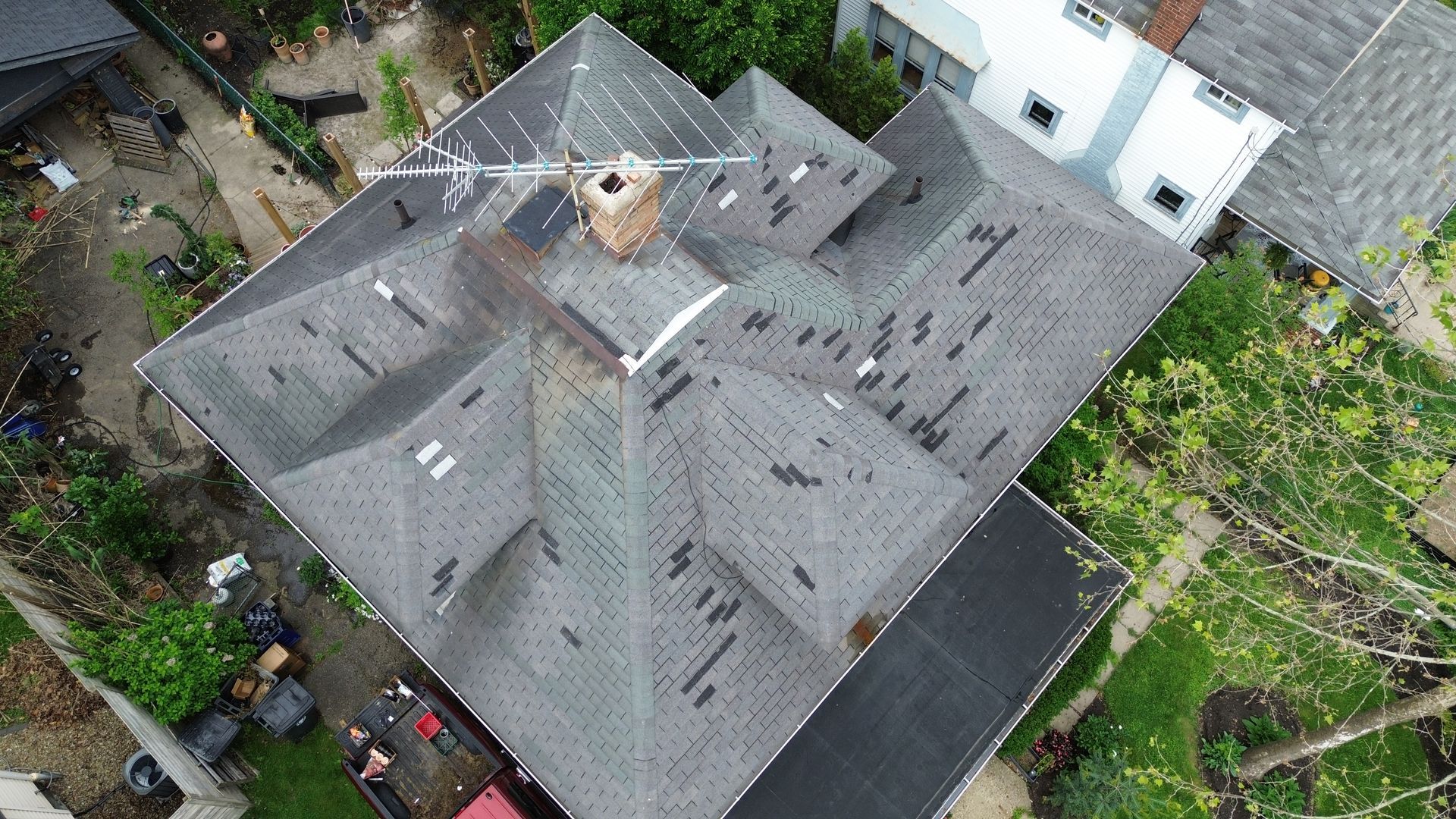 Aerial view of a gray roof with a central chimney, surrounded by trees and neighboring buildings.