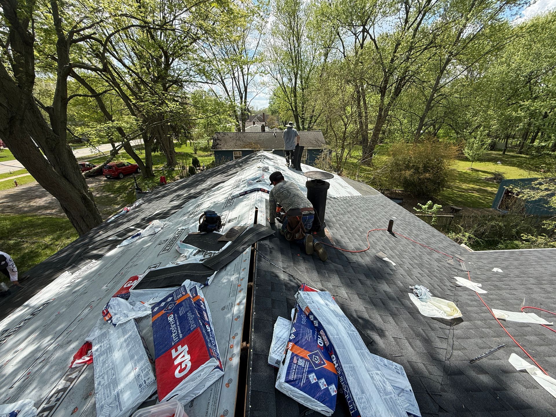 Roof covered in black tarp with tools and torn packaging, surrounded by trees in a yard