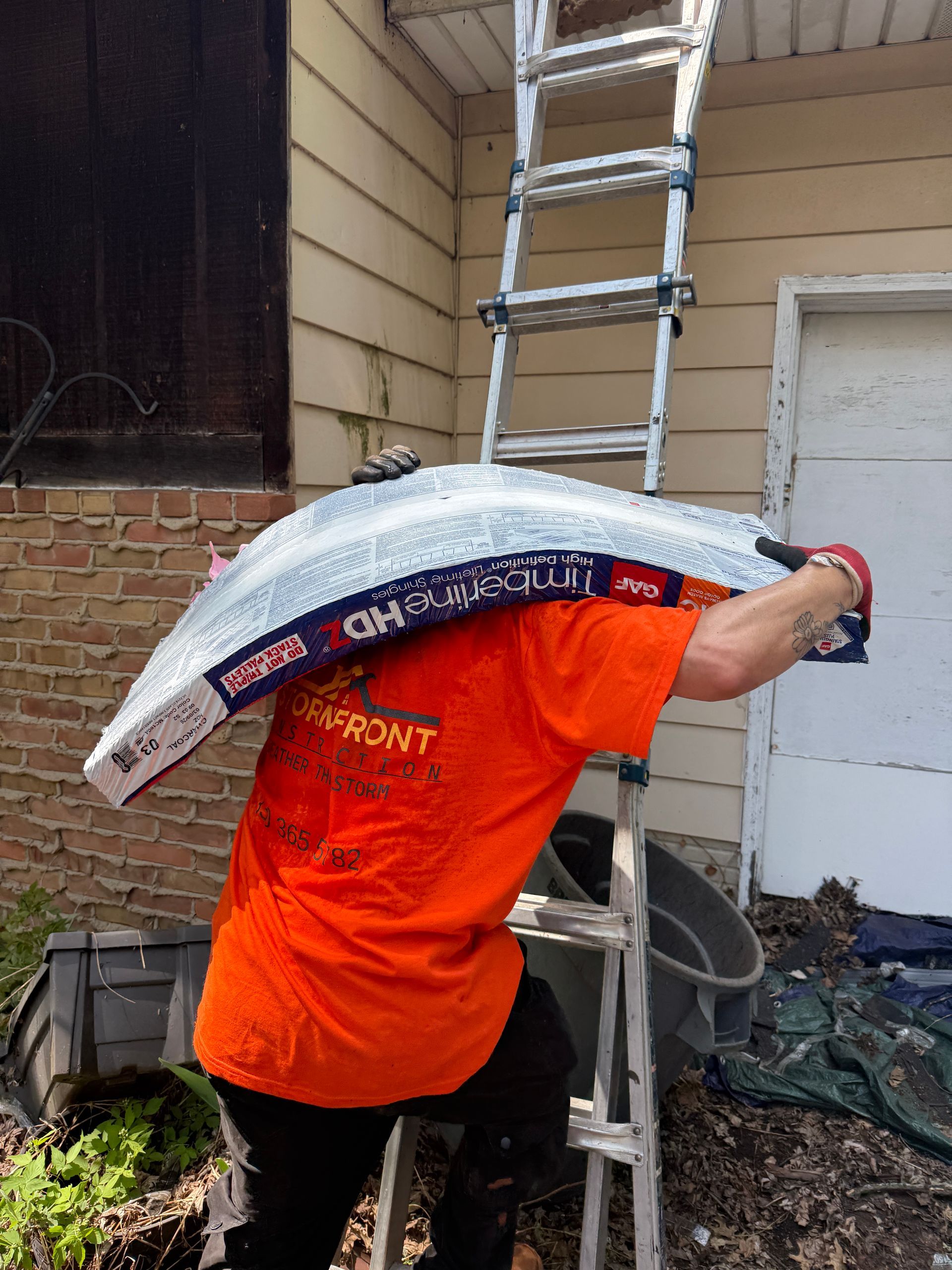 Worker in orange shirt carrying a ladder on a shoulder beside a house and brick wall