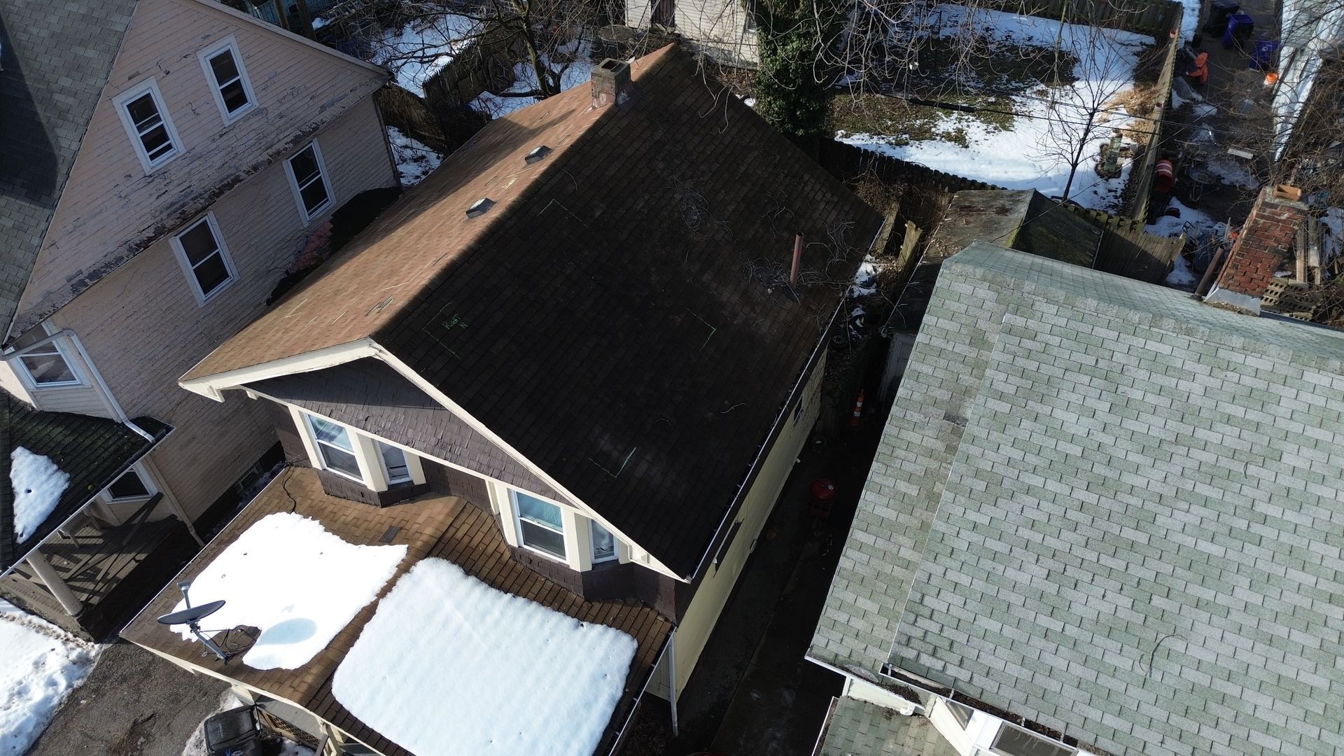 Snow-covered residential rooftops beside a house and gray roof in a winter neighborhood aerial view