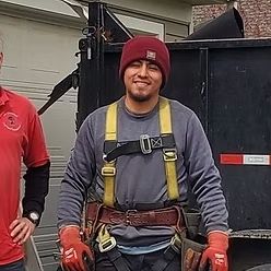 A smiling construction worker wearing a maroon beanie, grey sweatshirt, and yellow safety harness stands outdoors.