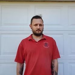 A person with a beard and tattoos on their arms wearing a red polo shirt, standing in front of a white garage door.
