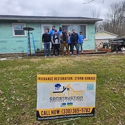 A construction crew stands before a house under renovation behind a Storm Front Construction sign in a grassy yard.
