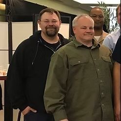 Three men stand close together indoors, wearing casual shirts and jackets, looking toward the camera.