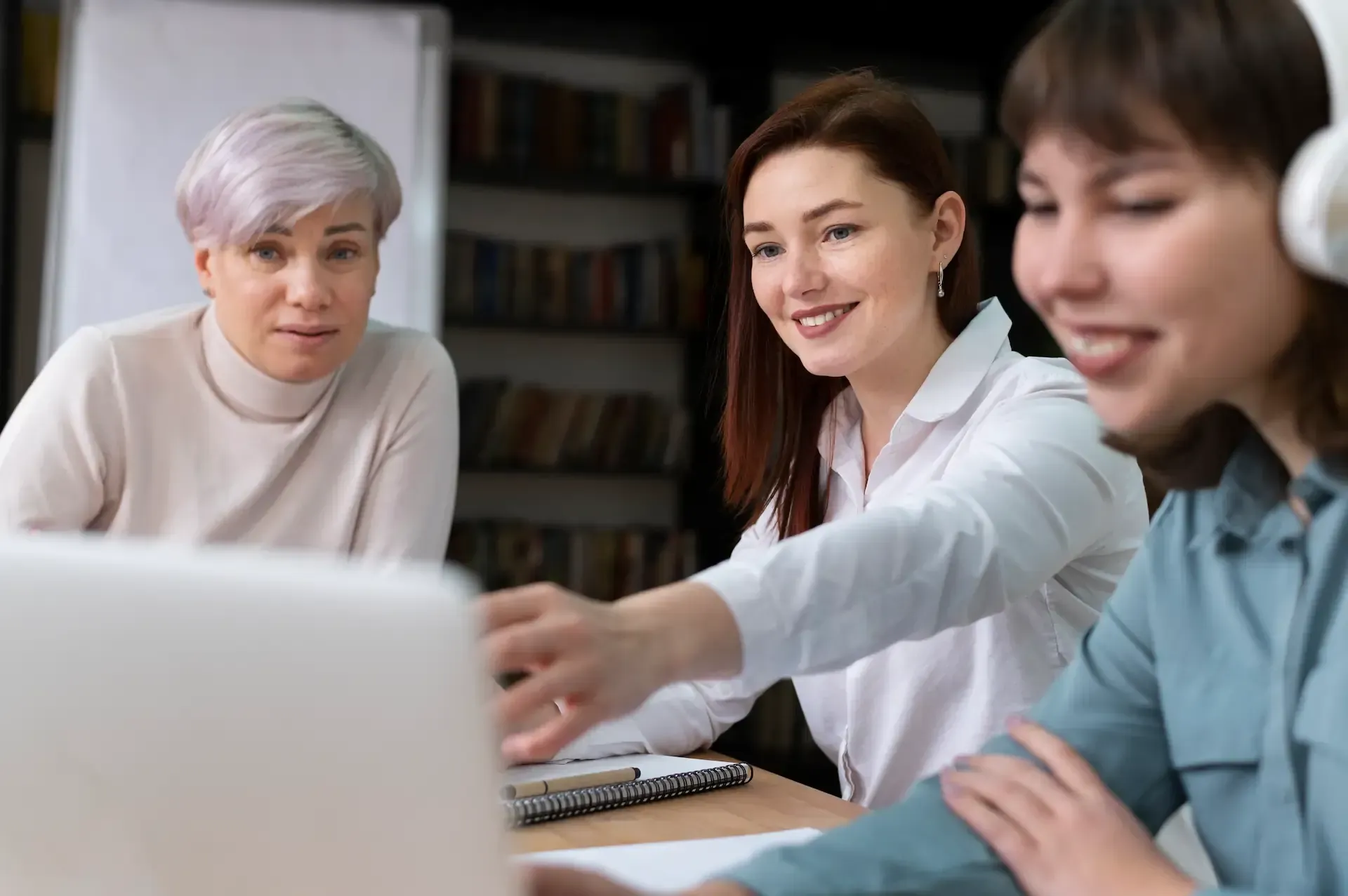 Three coworkers collaborating at a laptop in a bright office, one pointing at the screen.