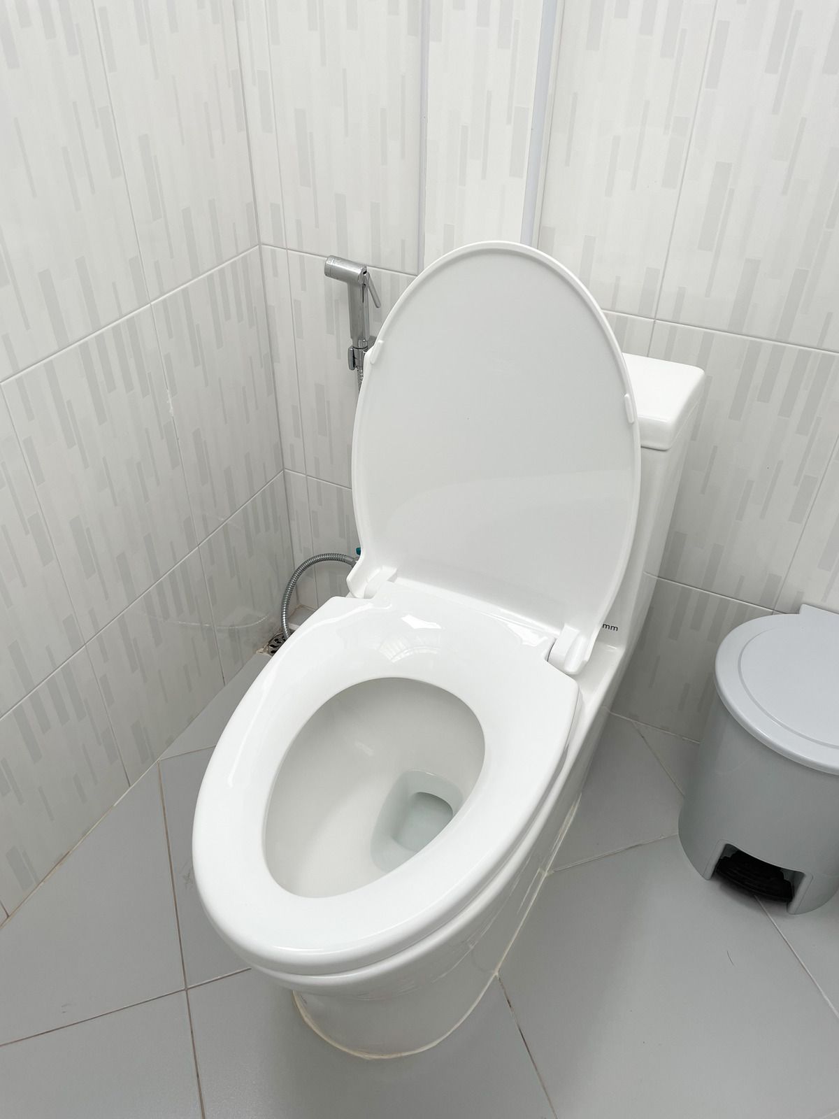 White toilet with seat up in a small tiled bathroom, with a toilet paper bin beside it.