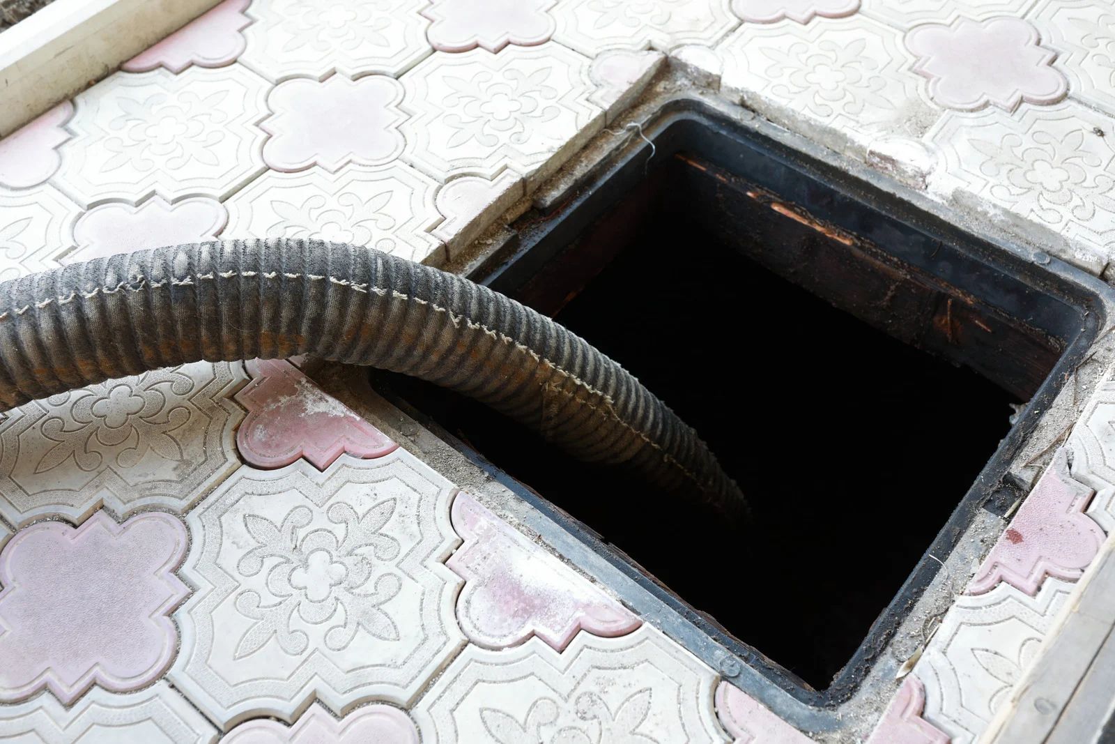 Hose inserted into an open, square manhole on a patterned pavement.