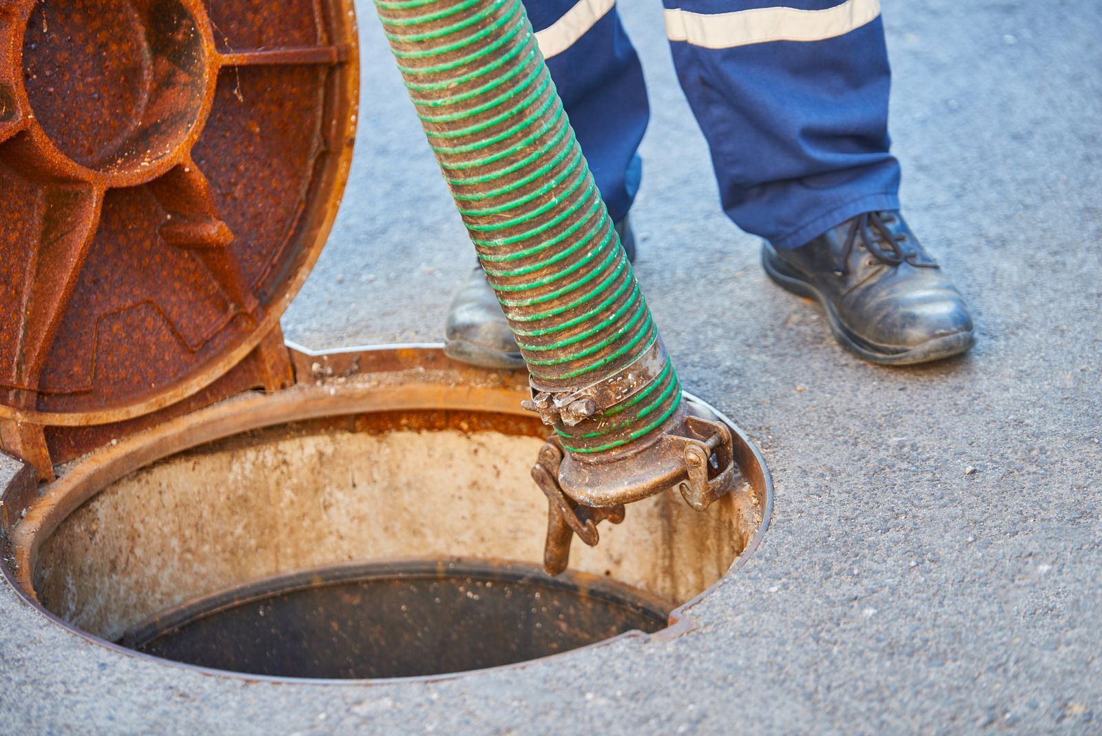 Worker using a green hose to clean a manhole beside a street, with the cover open nearby.