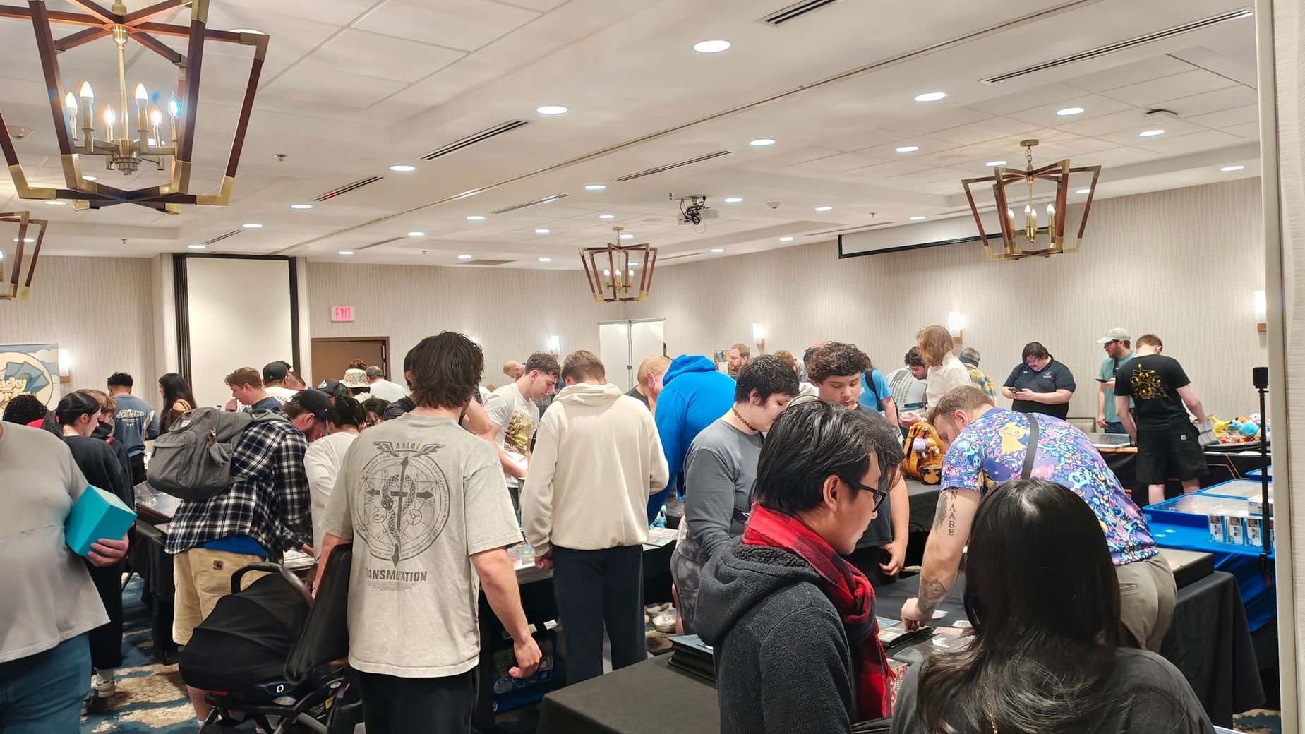 Crowded indoor market with people browsing tables of assorted items under chandeliers