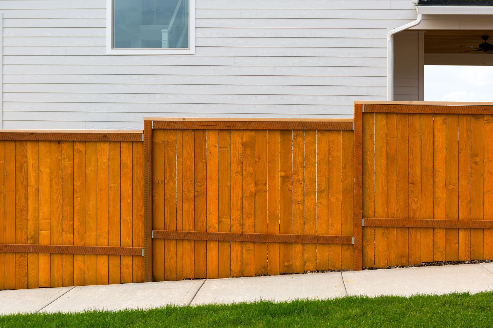 Wooden privacy fence in front of a white house, with green grass along the bottom. Wooden privacy fence in front of a white house, with green grass along the bottom.