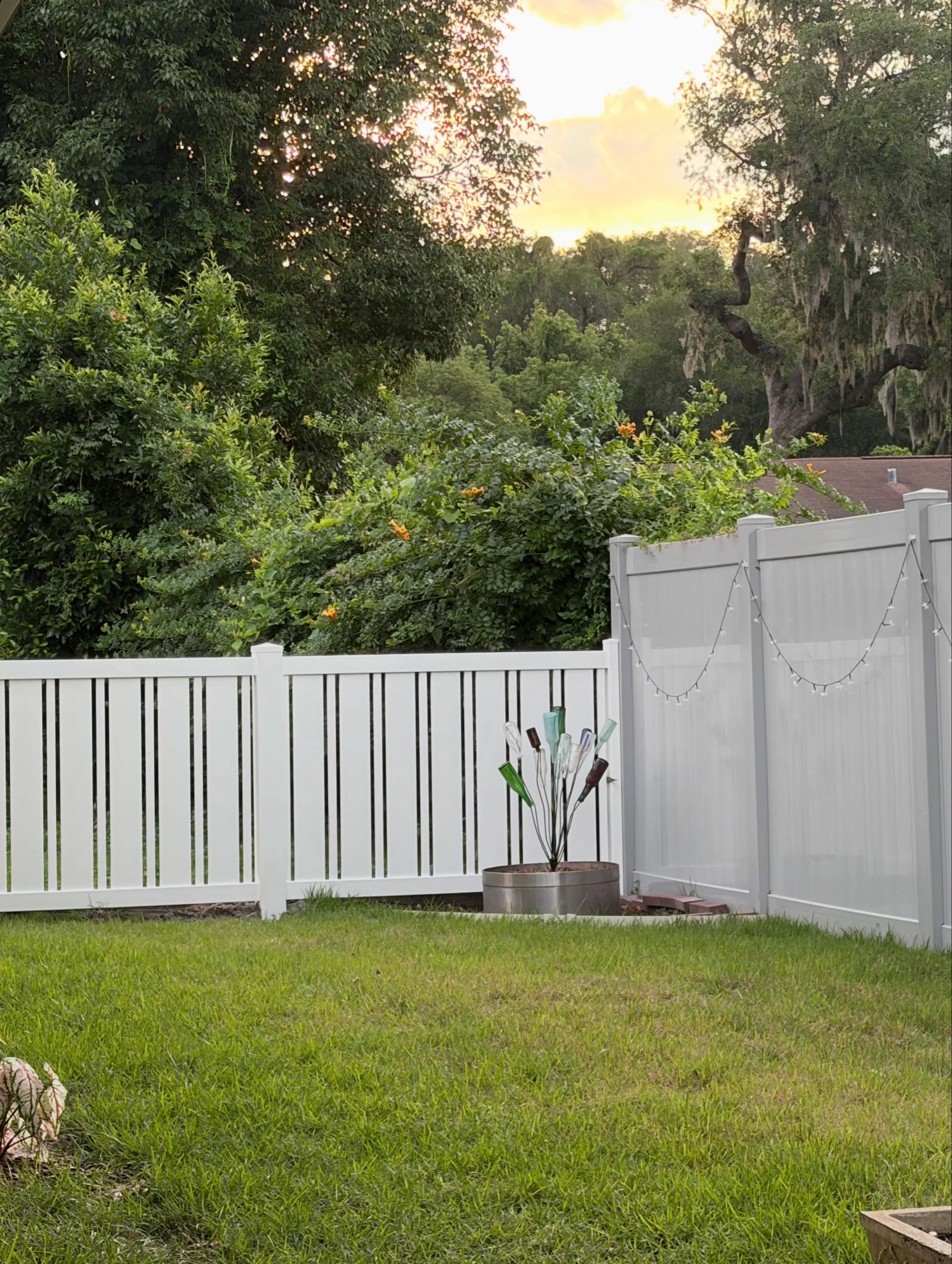 Wooden backyard fence beside a leafy yard and a house with a blue porch in the background Wooden backyard fence beside a leafy yard and a house with a blue porch in the background