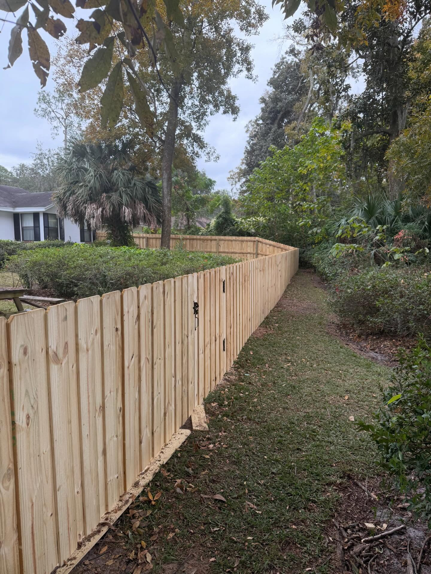 Wooden privacy fence along a grassy yard beside a gravel path, with a blue house in the background. Wooden privacy fence along a grassy yard beside a gravel path, with a blue house in the background.