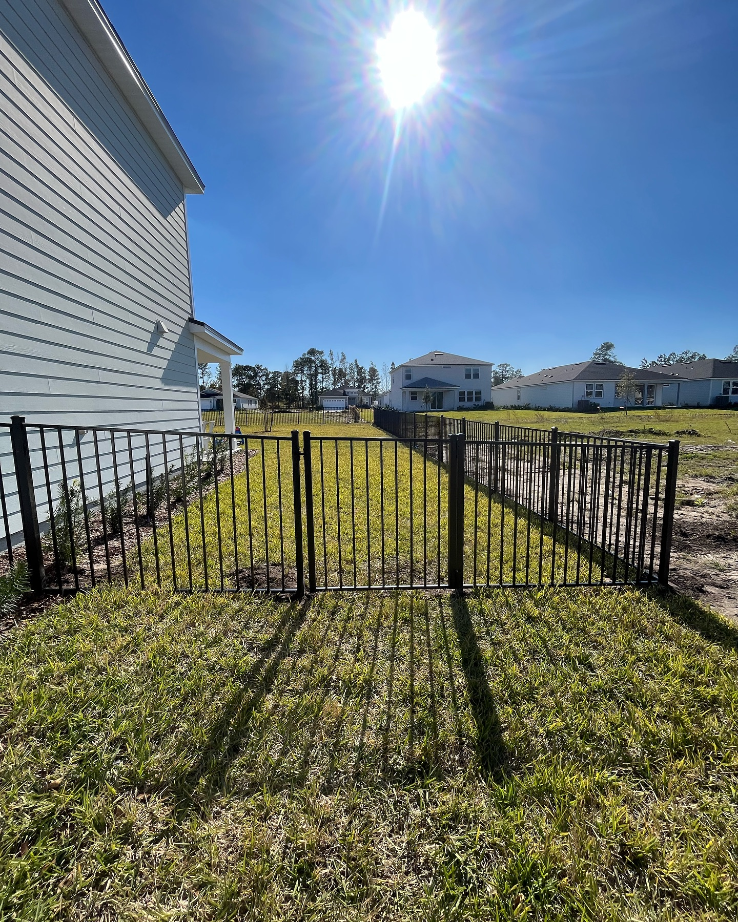 Black metal gate beside a white house, opening onto a grassy yard in bright sunlight Black metal gate beside a white house, opening onto a grassy yard in bright sunlight