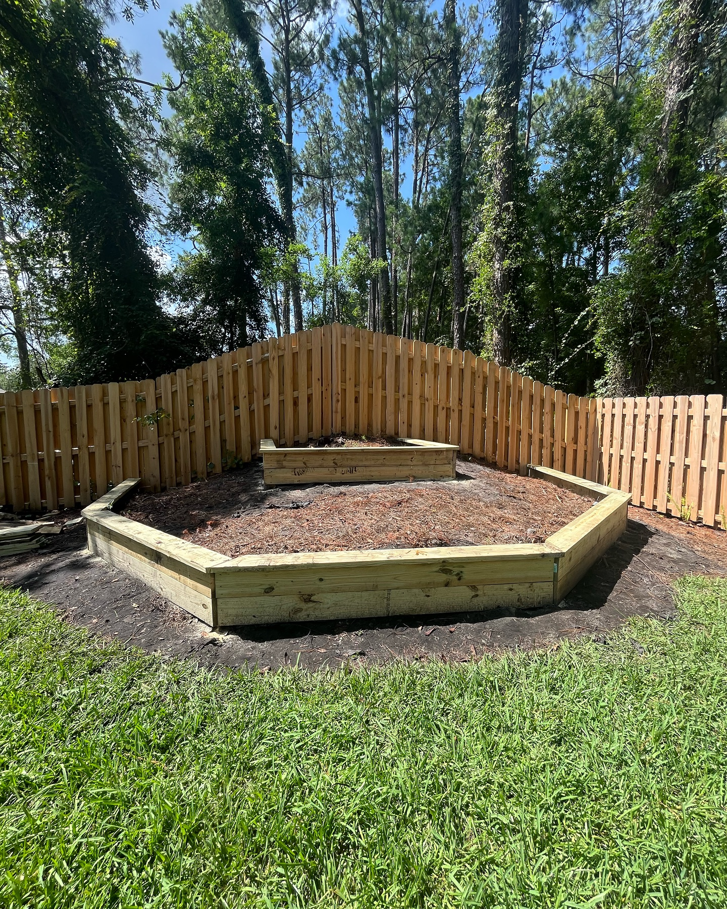 Wooden raised garden bed and fenced outdoor area in a sunny yard, surrounded by trees and grass Wooden raised garden bed and fenced outdoor area in a sunny yard, surrounded by trees and grass