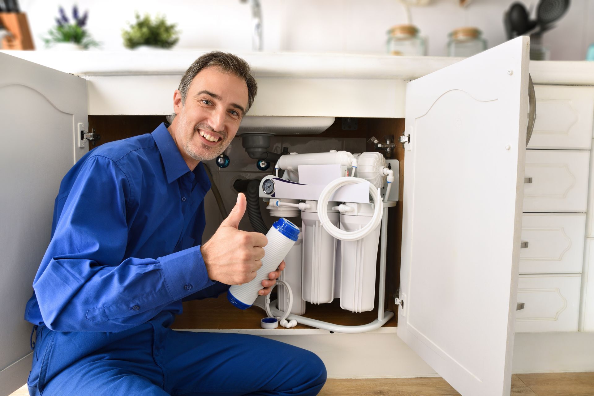 A man is kneeling under a sink holding a bottle and giving a thumbs up.