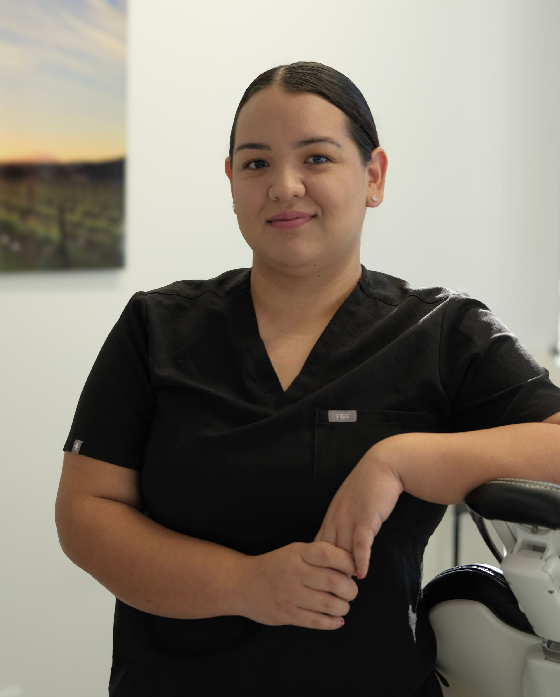 Woman in black scrubs, leaning on medical equipment, smiling in a clinical setting.
