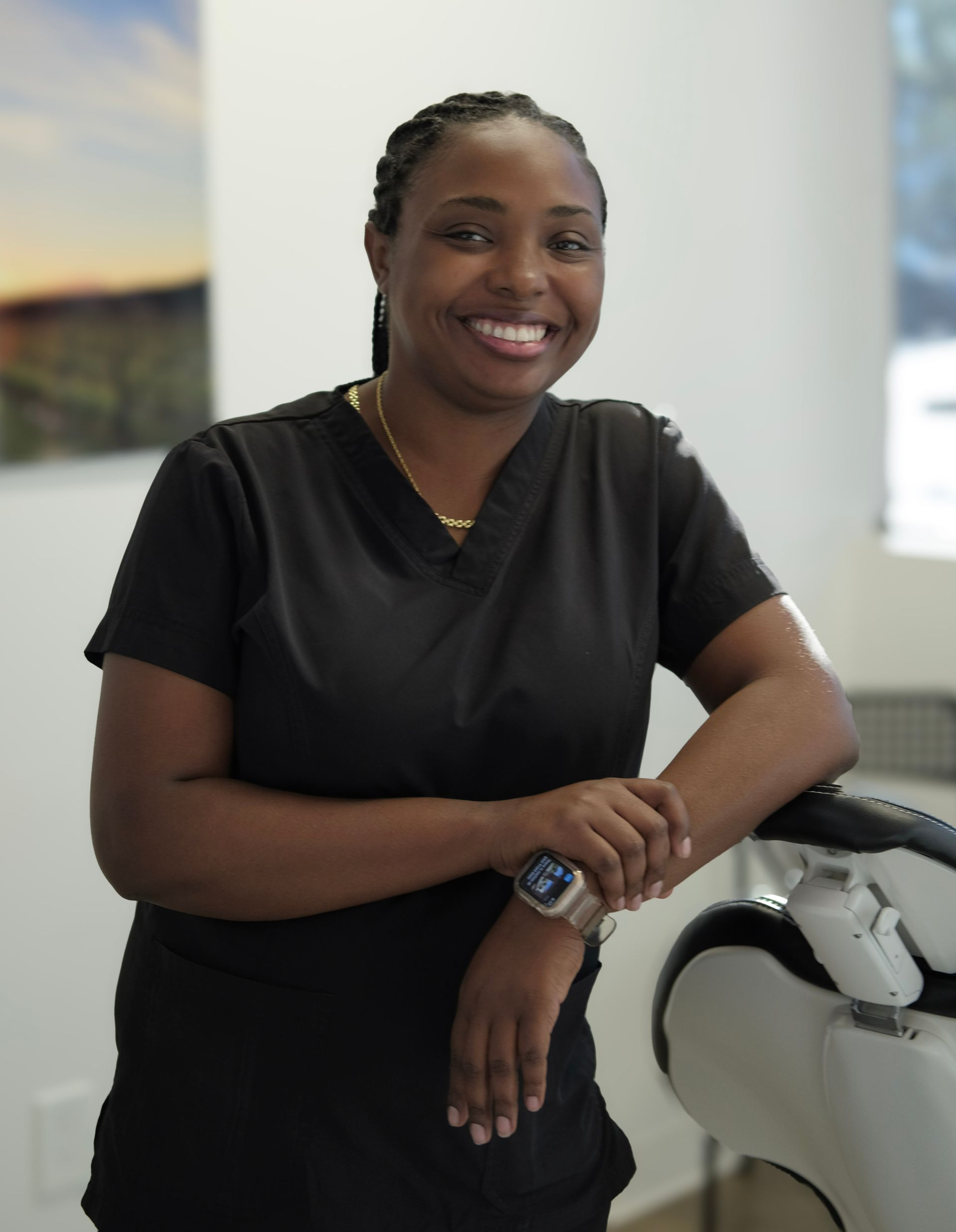 Smiling person in black scrubs, leaning on dental chair in a bright office.