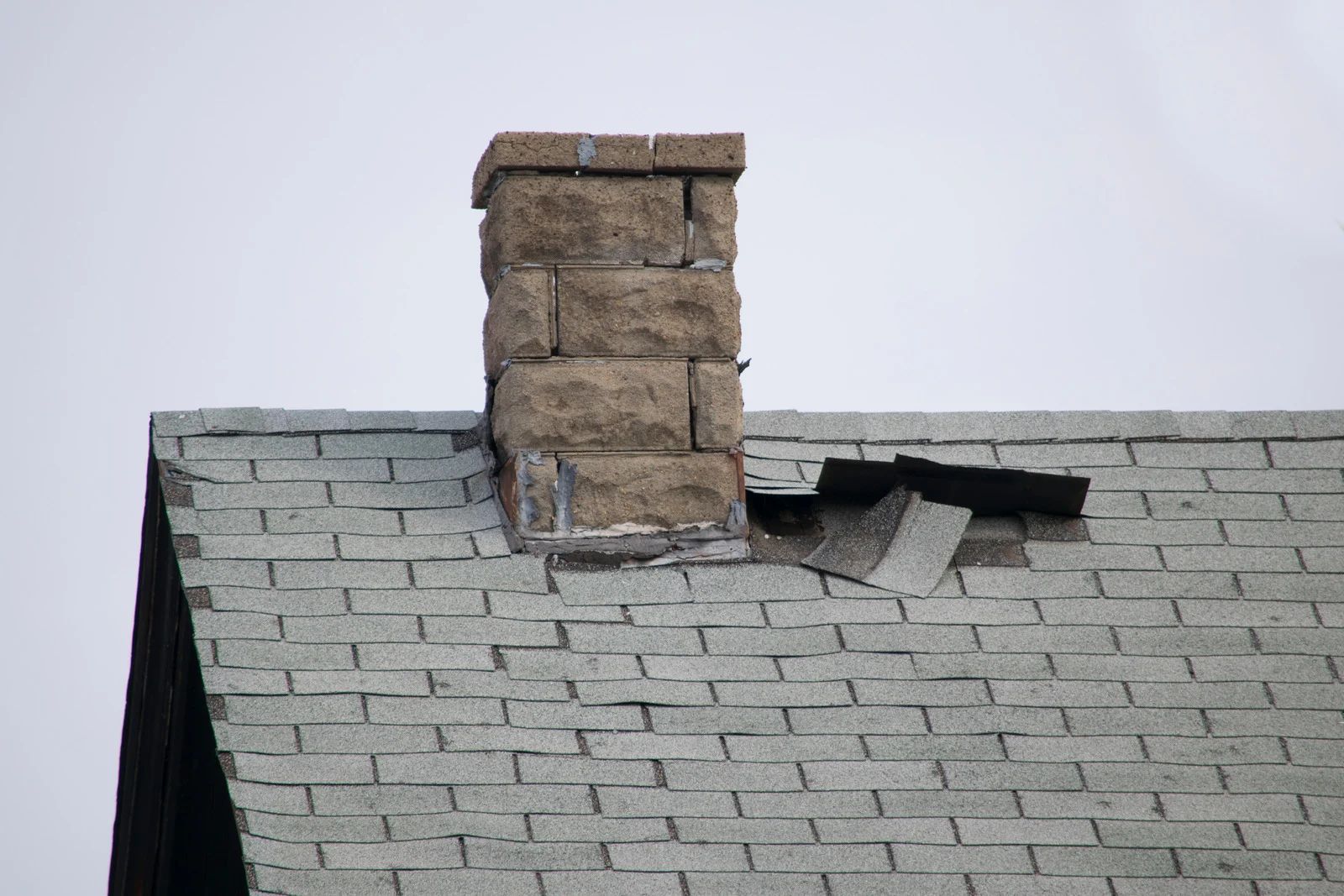 Damaged roof with a brick chimney and missing shingles on gray shingles against a pale sky