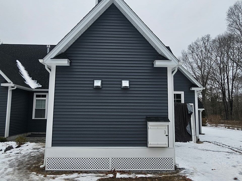 A blue-sided house exterior with two vents on the wall, a white utility box, and lattice skirting, set in snowy weather.