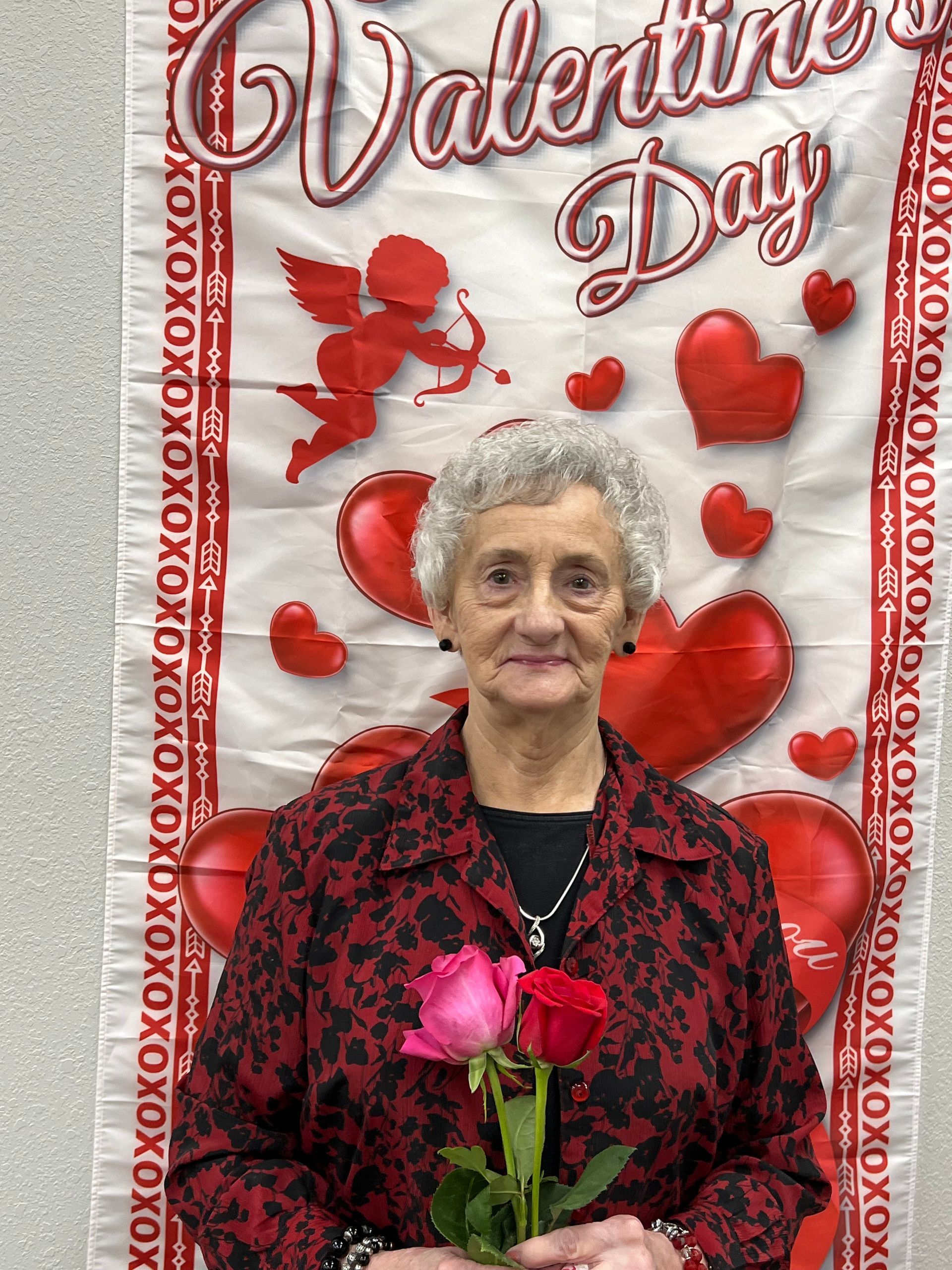 Woman holding roses in front of Valentine's Day banner, wearing a red patterned jacket.