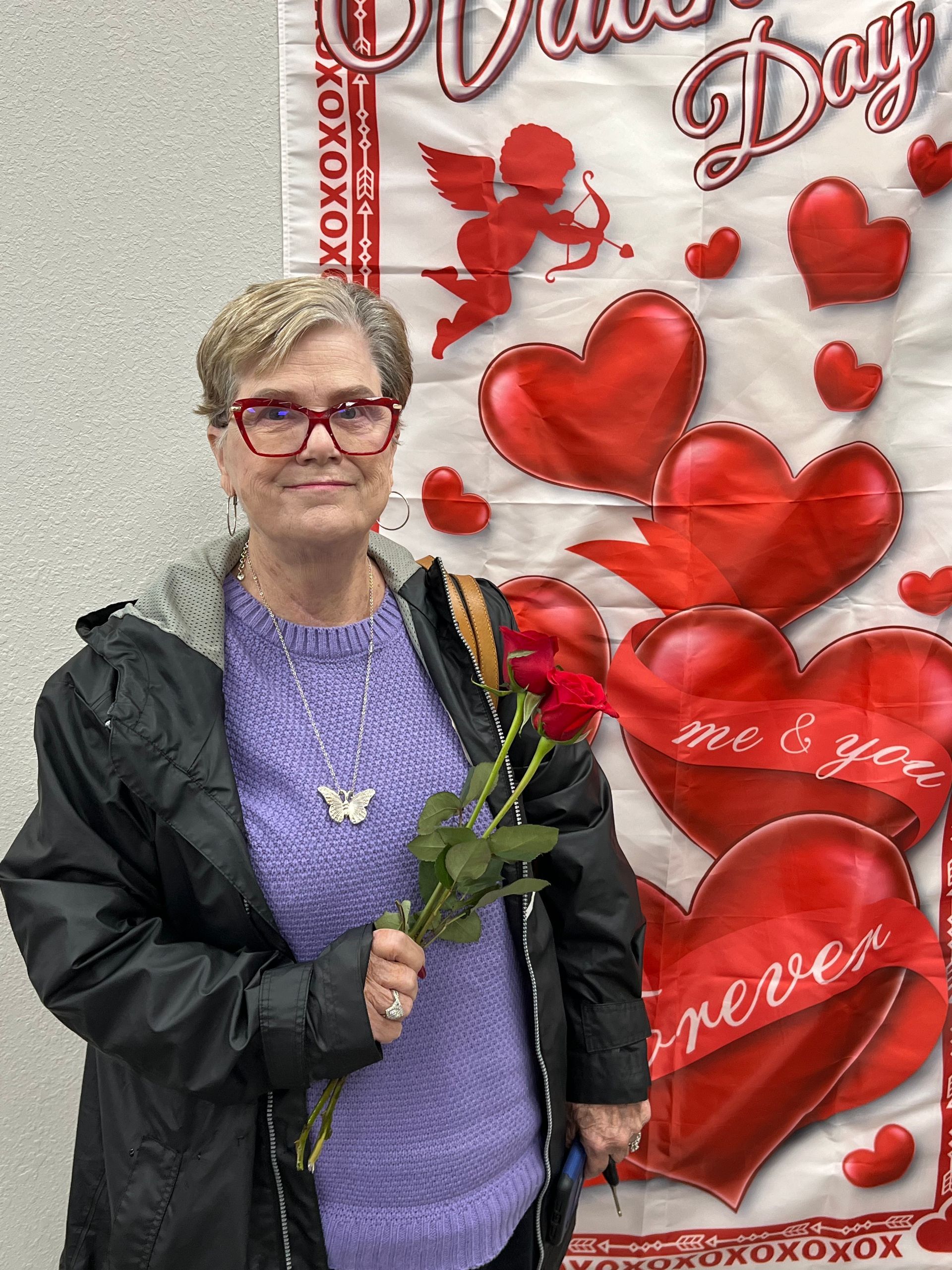 Woman holding roses stands in front of Valentine's Day banner with red hearts. She wears glasses and a black jacket.